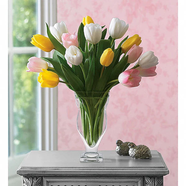Bouquet of yellow, pink, and white tulips in a clear vase on a decorative table with a pink wall background.