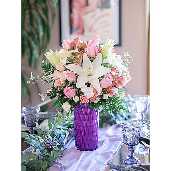 Floral arrangement in a purple vase on a table with glasses and greenery.
