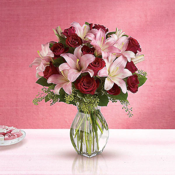 Bouquet of red roses and pink lilies in a clear vase on a pink background