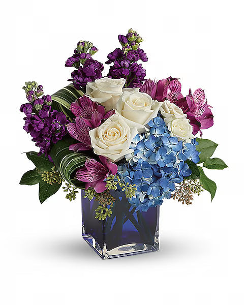 Floral arrangement with white roses, purple and pink blooms, and blue hydrangeas flowers in a clear vase on a white background