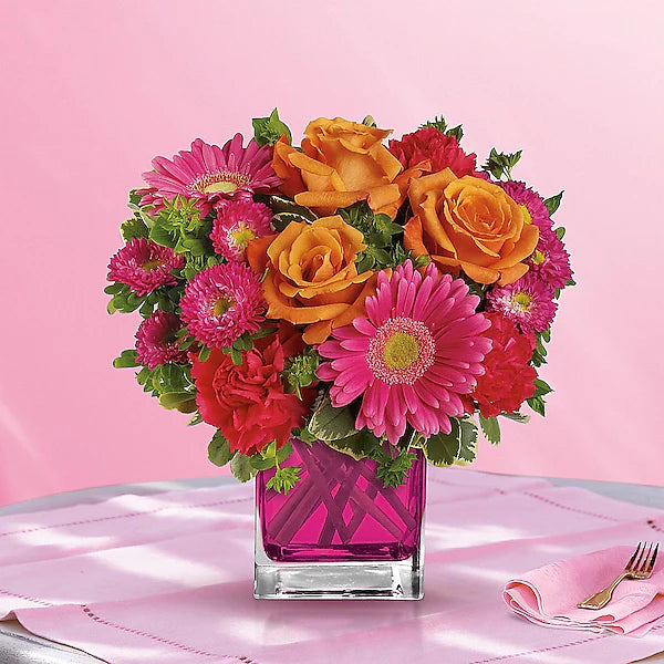 Colorful bouquet of flowers in a pink vase on a pink tablecloth with a fork and knife.