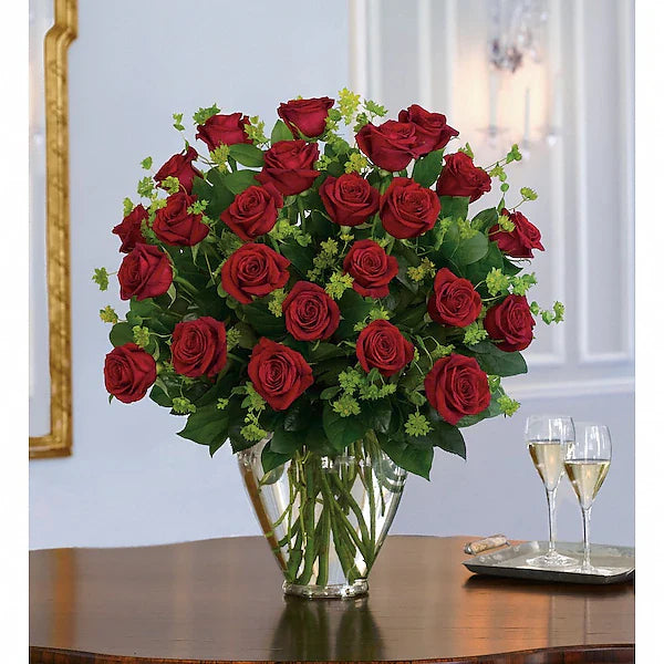 Bouquet of red roses in a clear vase on a table with two wine glasses.