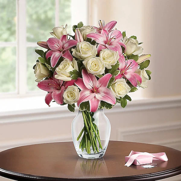 Bouquet of pink and white flowers in a clear vase on a wooden table with a blurred window background.