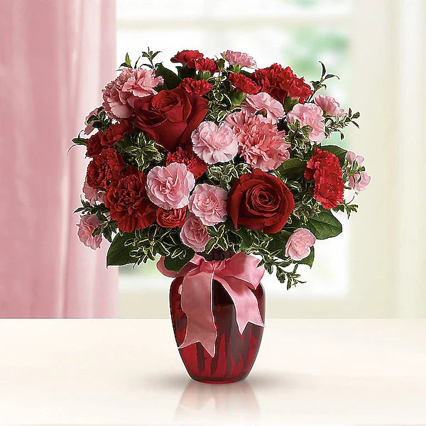 Colorful arrangement of red and pink flowers in a red vase, decorated with a pink ribbon