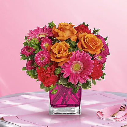 Colorful bouquet of flowers in a pink vase on a pink tablecloth with a fork and knife.