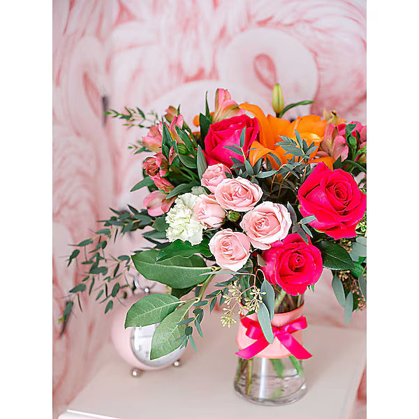Bouquet of mixed flowers in a clear glass vase tied with a pink ribbon against a pink flamingo-themed background.